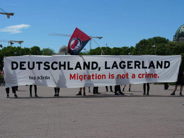 Eine Gruppe von Menschen hält eine Fahne und ein Transparent mit der Aufschrift 'Deutschland, Lagerland Migration ist kein Verbrechen' und steht auf dem Boden mit Straßenlaternen, Bäumen, Kränen, einem Gebäude und einem bewölkten Himmel im Hintergrund.