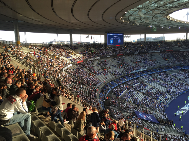 Große Menschenmenge in einem Stadion bei einem Fußballspiel mit einer Bühne, Fahnen, Stangen, einem Bildschirm und der Allianz Arena in München, Deutschland im Hintergrund.