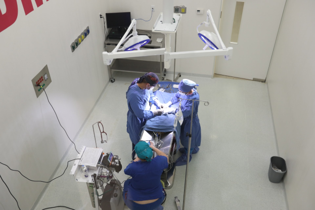 Chirurgen führen eine Operation an einem Patienten auf einer Trage in einem Operationssaal durch, mit medizinischer Ausrüstung an den Wänden und einem Objekt auf dem Boden rechts.