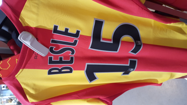 Ein roter und gelber Fußballtrikot mit Text und Zahlen hängt an einem Ständer in einem Geschäft, mit anderen Gegenständen und dem Boden im Hintergrund.