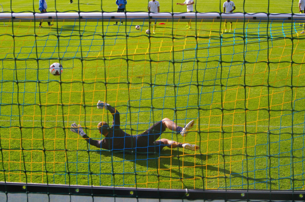 Ein Fußballtorhüter taucht ab, um einen Ball vor dem Tor zu halten, mit einem Netz im Vordergrund und einigen Menschen im Hintergrund, sowie mehreren Fußballen auf dem Boden verstreut.