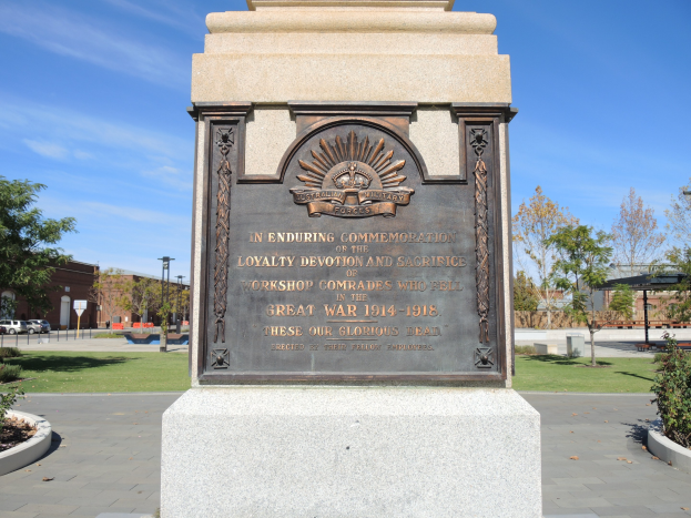Ein Denkmal mit einer Tafel, die 'Treue, Hingabe und Opfer' trägt, steht in einem Park, umgeben von Pflanzen, mit Bäumen, Gebäuden und Fahrzeugen im Hintergrund unter einem klaren blauen Himmel.