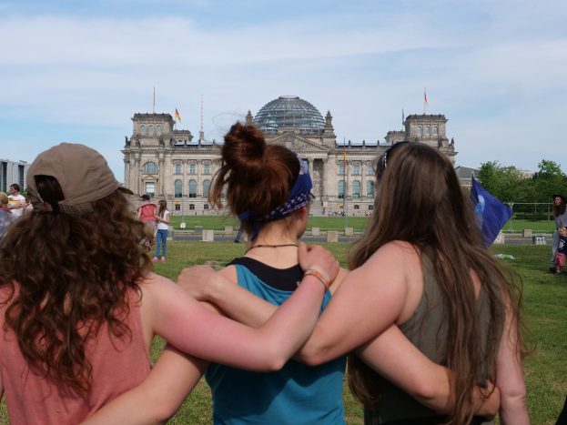 Drei Frauen in bunten Kleidern stehen vor dem Reichstag in Berlin, mit Menschen, Fahnen und Bäumen im Hintergrund.