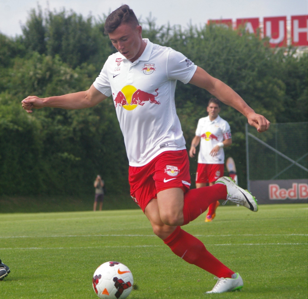 Ein professioneller Fußballspieler in einem roten und weißen Trikot kickt einen Fußball auf einem grünen Feld mit Bäumen und einem klaren blauen Himmel im Hintergrund, mit "RB Leipzig" auf einer Tafel dahinter.