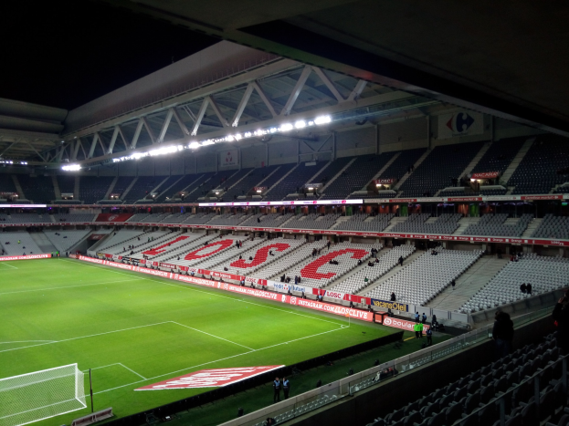 Großes Stadion mit einem Fußballfeld, das von grünem Gras bedeckt ist, einem Netz auf der linken Seite, ein paar Menschen auf den Tribünen und hellen Scheinwerfern an der Decke gegen einen dunklen Hintergrund.