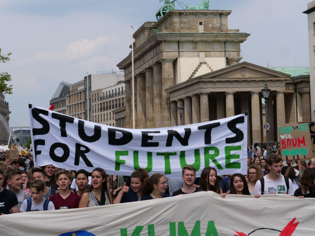 Gruppe von Schülern, die in Berlin mit einem bunten "Students for Future"-Schild marschieren, vor Gebäuden, Bäumen und Himmel.