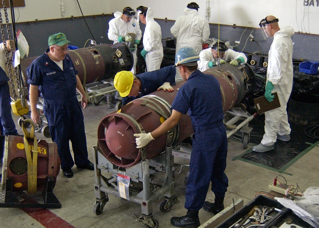 Männer in Helmen und Handschuhen, die eine große Bombe in einer Fabrik mit Maschinen und Wandtext auseinandernahmen.