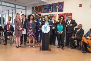 Eine Gruppe von Menschen steht vor einem Podium mit einem Mikrofon und einer Tafel mit der Aufschrift "Sancho Stadtverordneter tritt dem Stadtrat bei", während andere auf Stühlen sitzen oder in der Nähe stehen; Flaggen, ein Feuerlöscher und Deckenleuchten sind im Hintergrund zu sehen.