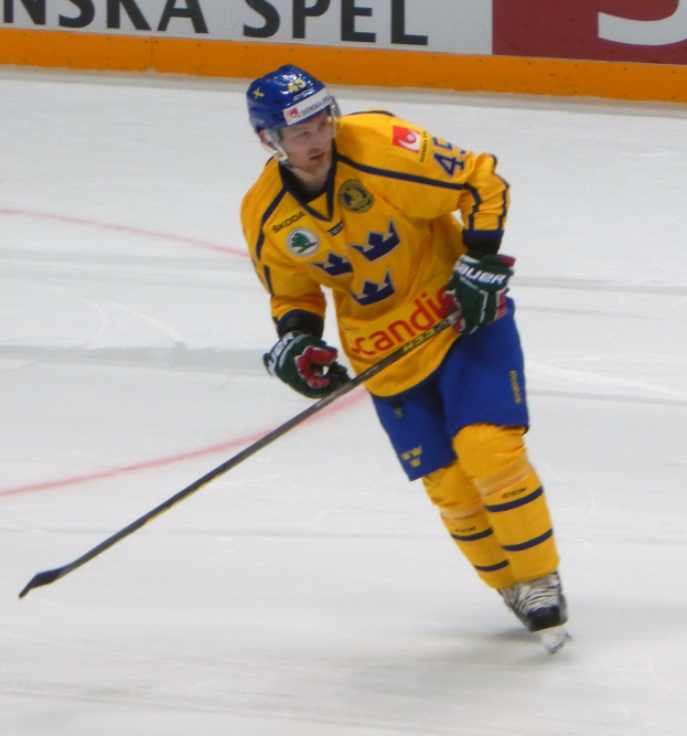 Ein Eishockeyspieler in Bewegung auf dem Eis mit Helm, Handschuhen und Schlittschuhen, der einen Hockeystock hält, mit einer Tafel im Hintergrund, auf der Text steht.