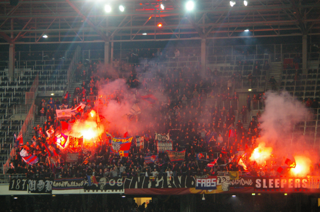 Eine große Menge Menschen in einem Stadion hält Fahnen und Banner, mit Leuchtraketen, die Rauch erzeugen, unter einem Dach mit Deckenlampen und Metallrahmen.