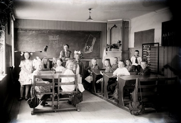 Schwarz-weißes Foto einer Klasse mit Kindern auf Bänken sitzend und stehend, mit Taschen, Wandtafel, Topfpflanzen, Schrank, Fenster und Deckenbeleuchtung.