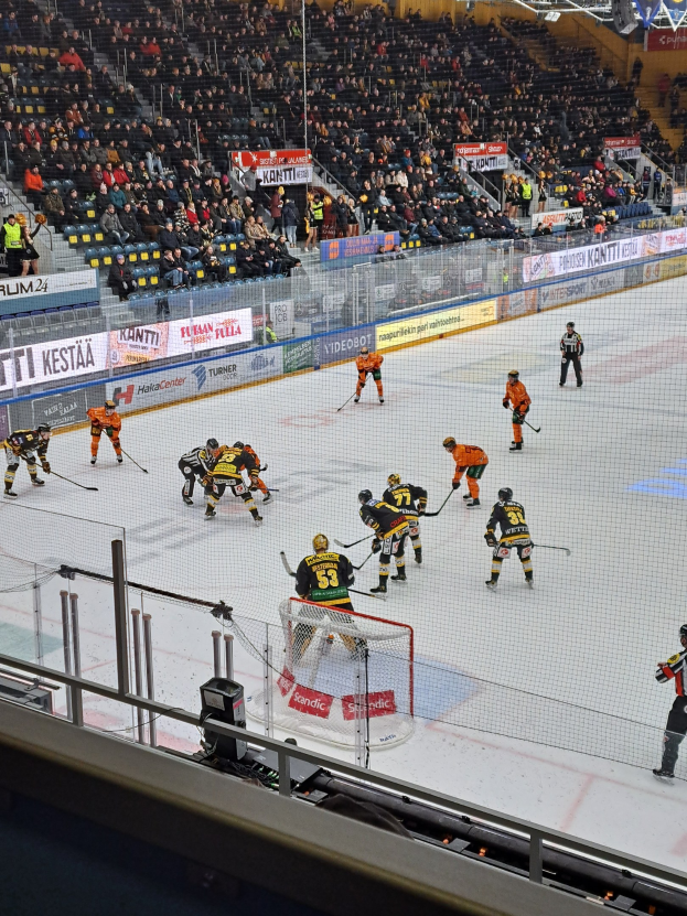 Eine Gruppe von Menschen, die Hockey auf einem Eisplatz spielen, umgeben von einem Zaun, mit Zuschauern im Hintergrund und beleuchteten Bannern darüber.