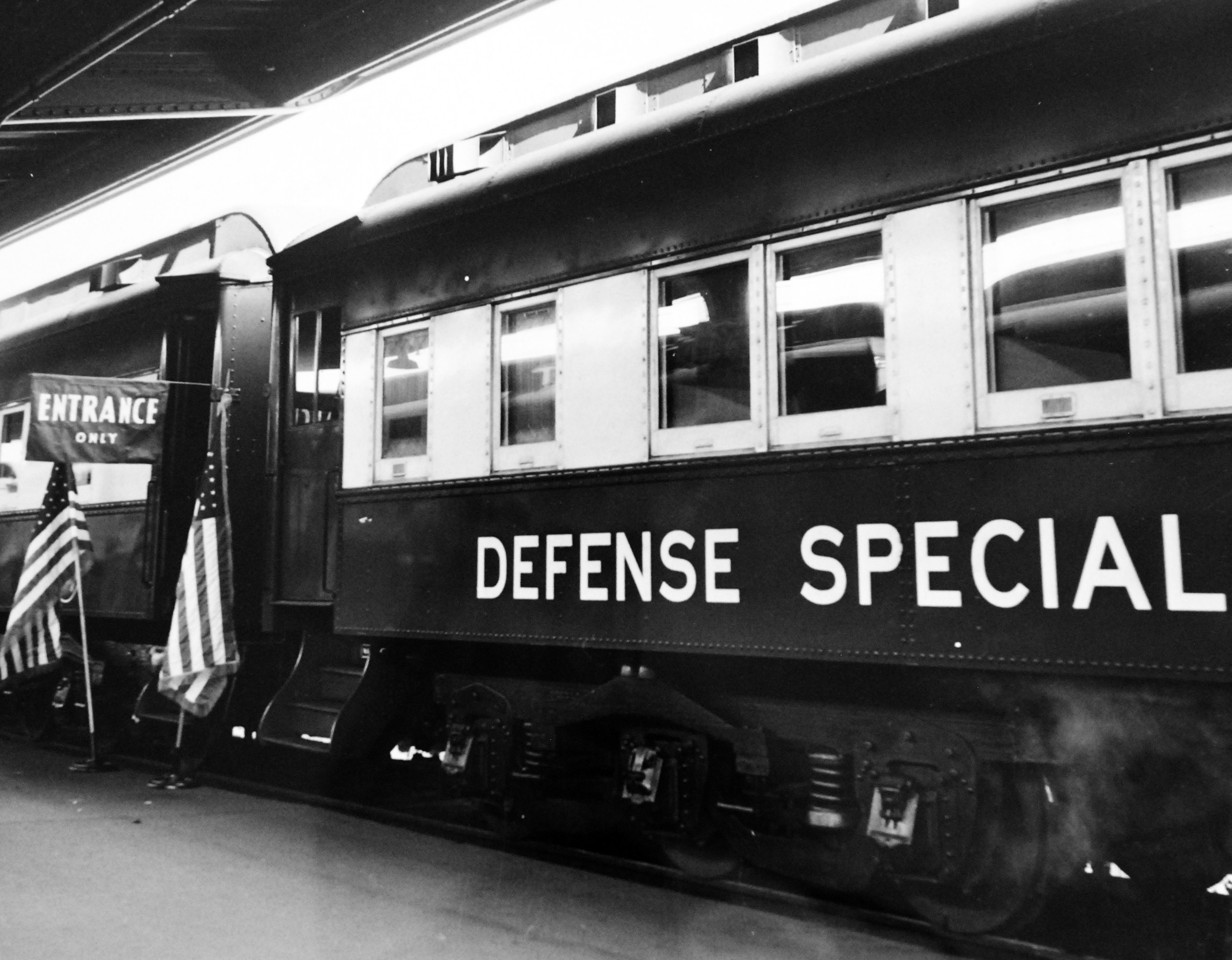 Schwarz-weiß-Foto eines Zuges mit der Aufschrift "Defense Special" auf den Eisenbahnschienen mit zwei Flaggen links und einer Decke oben.