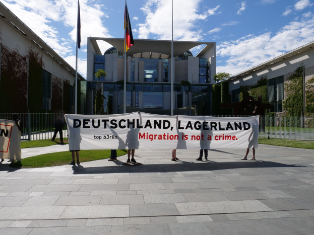 Eine Gruppe von Menschen hält ein Banner mit der Aufschrift "Deutschland, Lagerland Migration ist kein Verbrechen" vor einem Gebäude mit Fenstern, umgeben von Gras, einem Metallzaun, Pflanzen, Bäumen und Fahnen mit Stangen unter einem bewölkten Himmel.