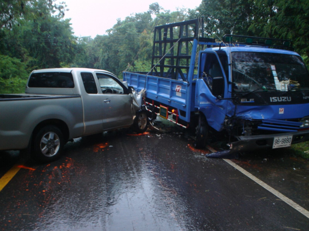 Ein schwer beschädigtter Lkw mit eingedellter Front und verbeulter Karosserie steht neben der Straße, umgeben von Bäumen unter einem klaren blauen Himmel.