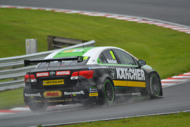 Ein Auto mit Text an der Seite fährt auf einer regennassen Rennstrecke, die von Gras gesäumt ist, mit einem Geländer und einem hölzernen Gebäude im Hintergrund.