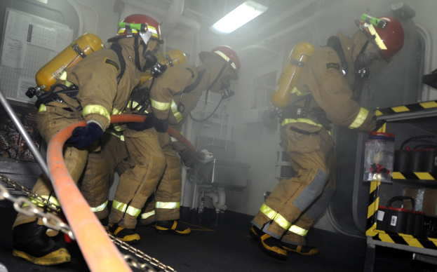 Feuerwehrleute in Schutzausrüstung, einschließlich Helmen und Sauerstoffflaschen, arbeiten an einem Löschfahrzeug mit Gestell von Gegenständen und Hintergrundrohren und -leuchten.