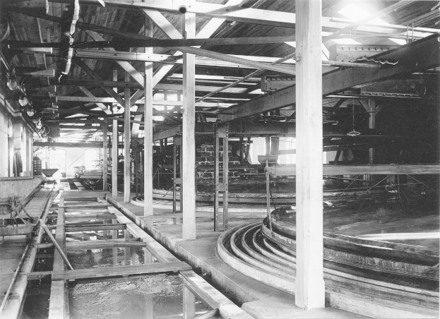 Schwarzes und weißes Foto einer Fabrik mit Pfählen, Lampen und anderen Gegenständen im Vordergrund, mit einem Wasserzeichen in der rechten unteren Ecke.