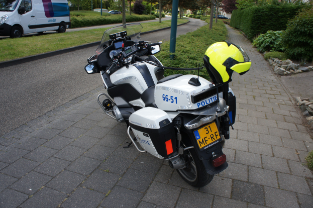 Ein Polizeimotorrad steht am Straßenrand mit einem Helm darauf, umgeben von Bäumen, Gebäuden und anderen Fahrzeugen.