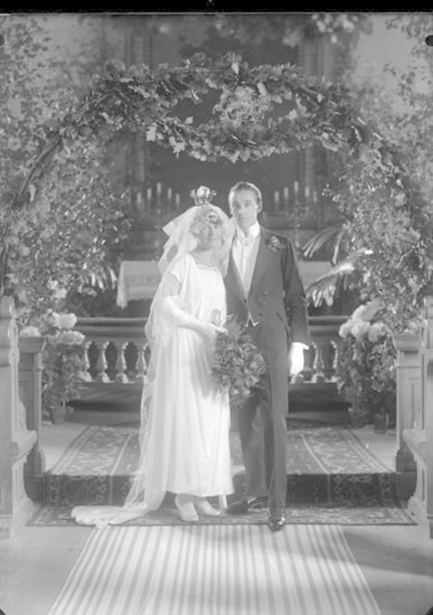 Schwarz-weiß-Foto eines Mannes und einer Frau, die unter einem Blumenschmuckbogen stehen, die Frau hält einen Strauß Blumen.