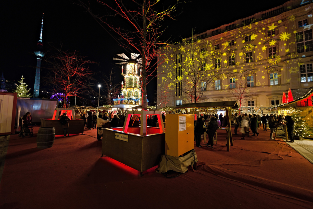 Ein geschäftiges Weihnachtsmarkt in Berlin, Deutschland mit Menschen um geschmückte Stände, festliche Lichter, Bäume, Gebäude, Laternenmäste und einen Turm unter einem dunklen Himmel.