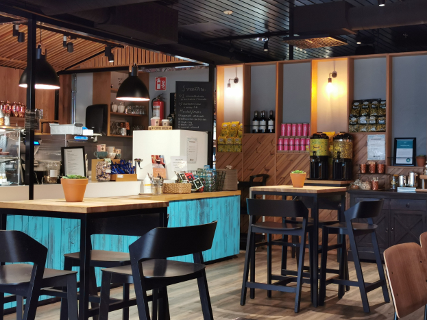 Innenraum eines Restaurants mit Holztischen, Stühlen, einer Topfpflanze auf einem Tisch, einer Tafel, einem Kühlschrank, einem Feuerlöscher, Regalen mit Flaschen, Schränken mit Gegenständen, einem Foto auf der Wand, Deckenlampen und Lampen.