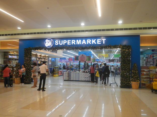 Eine Gruppe von Menschen, die vor einem Supermarkt stehen, einige halten Einkaufstäsche, mit einem Einkaufswagen, Topfpflanze, Regalen, Schildern und Deckenbeleuchtung im Hintergrund.