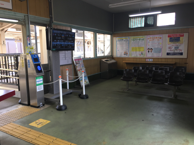Ein belebter Bahnhofseingang mit Reihen von Stühlen, einem Service-Schalter, Ticketautomaten, Deckenleuchten, Geländern, Informationsanzeigen und Wandplakaten.