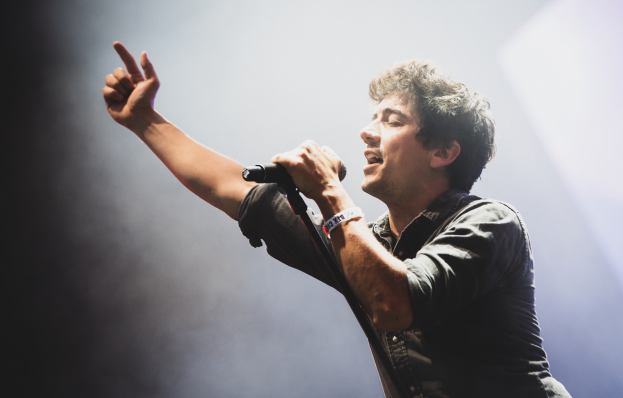 Ein Mann singt in ein Mikrofon auf einer hell erleuchteten Bühne, trägt ein schwarzes Hemd und Jeans und hat seine Haare wild gestylt, während das Publikum applaudiert.