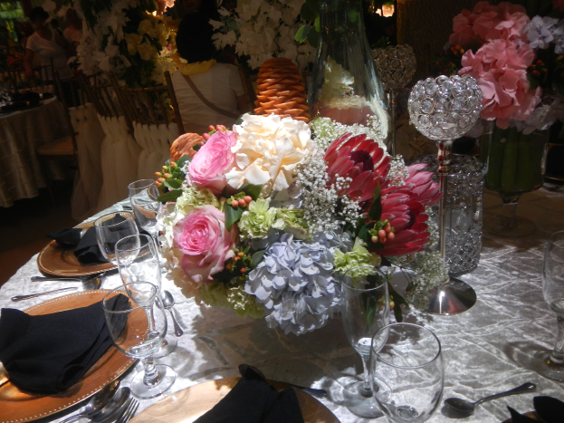 Ein formeller Esstisch mit Tellern, Besteck, Gläsern, Servietten und Blumenvasen, mit einem floralen Zentrum und Stühlen und besetzten Tischen im Hintergrund.