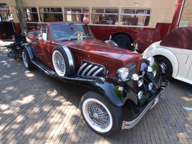 Vintage-Auto neben einem Doppeldeckerbus auf einer Kopfsteinpflasterstraße geparkt, mit Stühlen und einem Baum im Vordergrund, einem Gebäude mit Fenstern im Hintergrund und Menschen im Bus zu sehen.