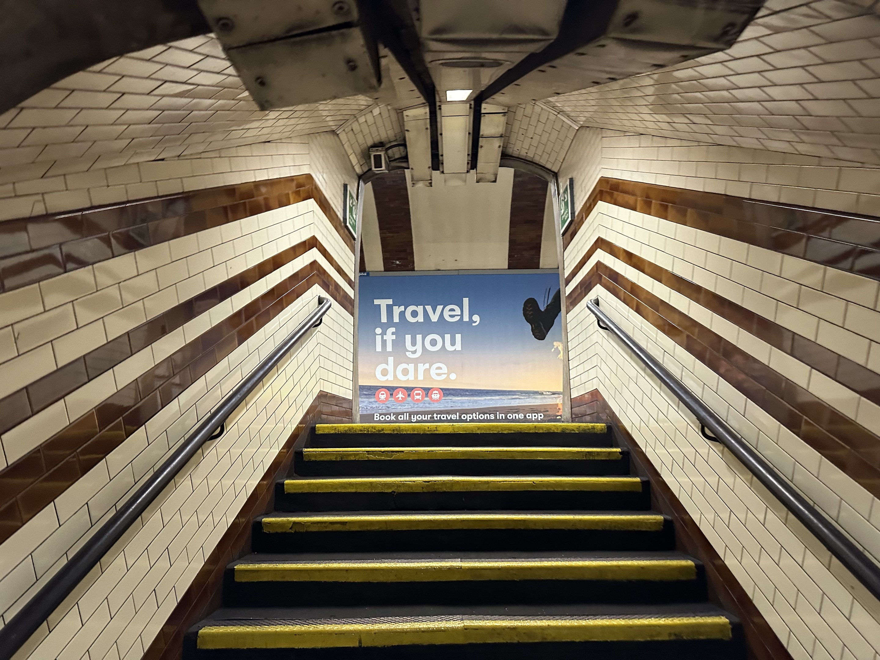 Eine Treppe in einer U-Bahn-Station mit einem "Reise, wenn du wagst"-Schild oben, flankiert von Geländern und Stationwänden im Hintergrund.