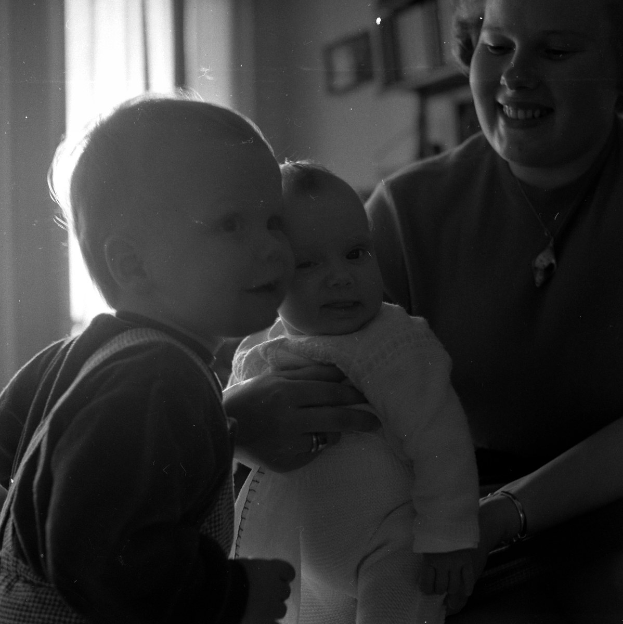 Eine Frau hält ein Baby mit einem warmen Lächeln in den Armen, während zwei andere Kinder neben ihr stehen, in einem gemütlichen Raum mit Rahmen an der Wand und einem Fenster im Hintergrund.