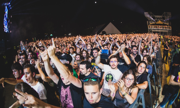 Eine große Menschenmenge auf einem Musikfestival mit erhobenen Händen, sitzende und stehende Besucher, ein Metallzaun auf der rechten Seite, Gebäude und Lichter im Hintergrund und ein dunkler Himmel, mit einem Mann und einer Frau im Vordergrund.
