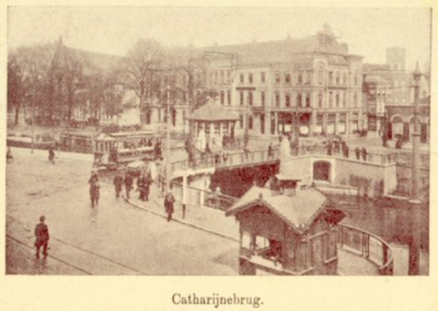 Schwarz-weißes Foto einer Stadtstraße mit Passanten, Gebäuden, Bäumen, Strommasten und einer Brücke im Hintergrund, mit Text unten.