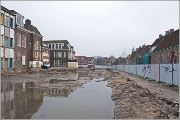 Eine überflutete Straßen in einer Stadt mit Wasser auf dem Boden, einem Zaun auf der rechten Seite, Gebäuden mit Fenstern auf der linken Seite, Bäumen im Hintergrund und einem Himmel darüber.