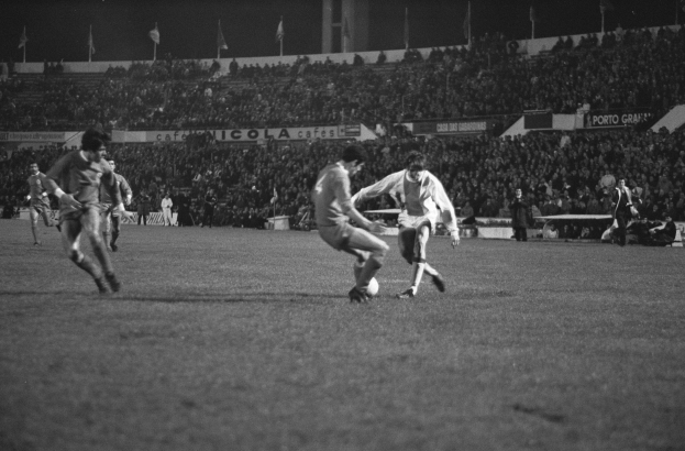 Schwarzes und weißes Foto von Männern, die Fußball auf einem Feld mit Zuschauern in den Rängen und Fahnen im Hintergrund spielen.