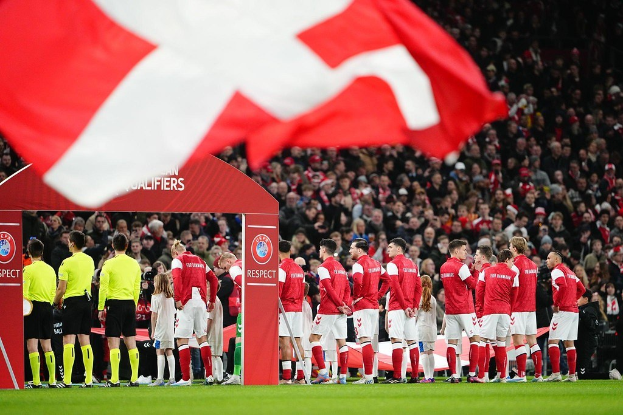 Gruppe von Menschen auf einem Fuballfeld mit einer roten und weißen Flagge im Vordergrund, einem Bogen mit 'Bayern Munich vs Bayern Munich Betting Tips & Preview' im Hintergrund und einer großen Menge im Stadion.