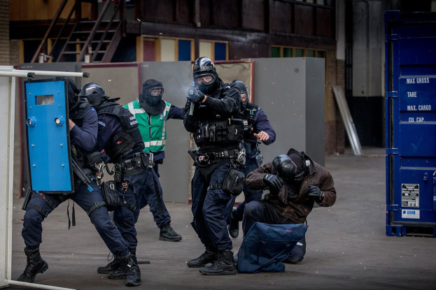 Polizeibeamte in Einsatzausrüstung stehen vor einem Gebäude, einige halten Waffen, mit einer Person auf dem Boden im Vordergrund und einem blauen Objekt mit Text auf der rechten Seite.