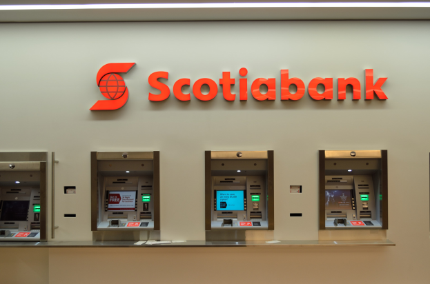 Ein modernes Scotiabank-Geldautomatengerät mit schwarzer und silberner Gestaltung, das eine große Anzeigefläche und Steuerungsknöpfe aufweist und vor einer hellgrauen Wand mit gerahmten Bildern und dem Text "Schottische Bank" im Hintergrund positioniert ist.