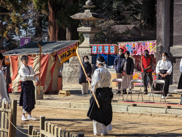 Eine Gruppe von Menschen in formeller Kleidung steht draußen in Kyoto, einige tragen Masken und halten hölzerne Stäbe, umgeben von einem Gebäude mit Bannern, einem Zelt und einem hölzernen Zaun unter einem klaren blauen Himmel.