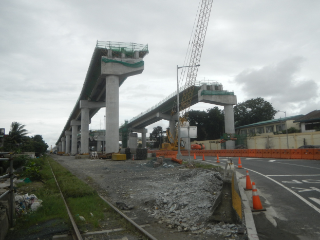 Baustelle mit einer Brücke im Hintergrund, Straße mit Absperrbaken markiert, Bahnschiene auf der linken Seite, verstreute Steine und Gras, Bäume und Gebäude auf beiden Seiten der Straße und ein bewölkter Himmel.