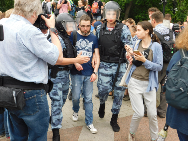 Eine Gruppe von Menschen steht vor einer größeren Menge, einige tragen Helme und Taschen, andere halten Kameras und Telefone; Einsatzpolizisten sind im Hintergrund bei Bäumen und Laternenpfählen in einem öffentlichen Bereich zu sehen.