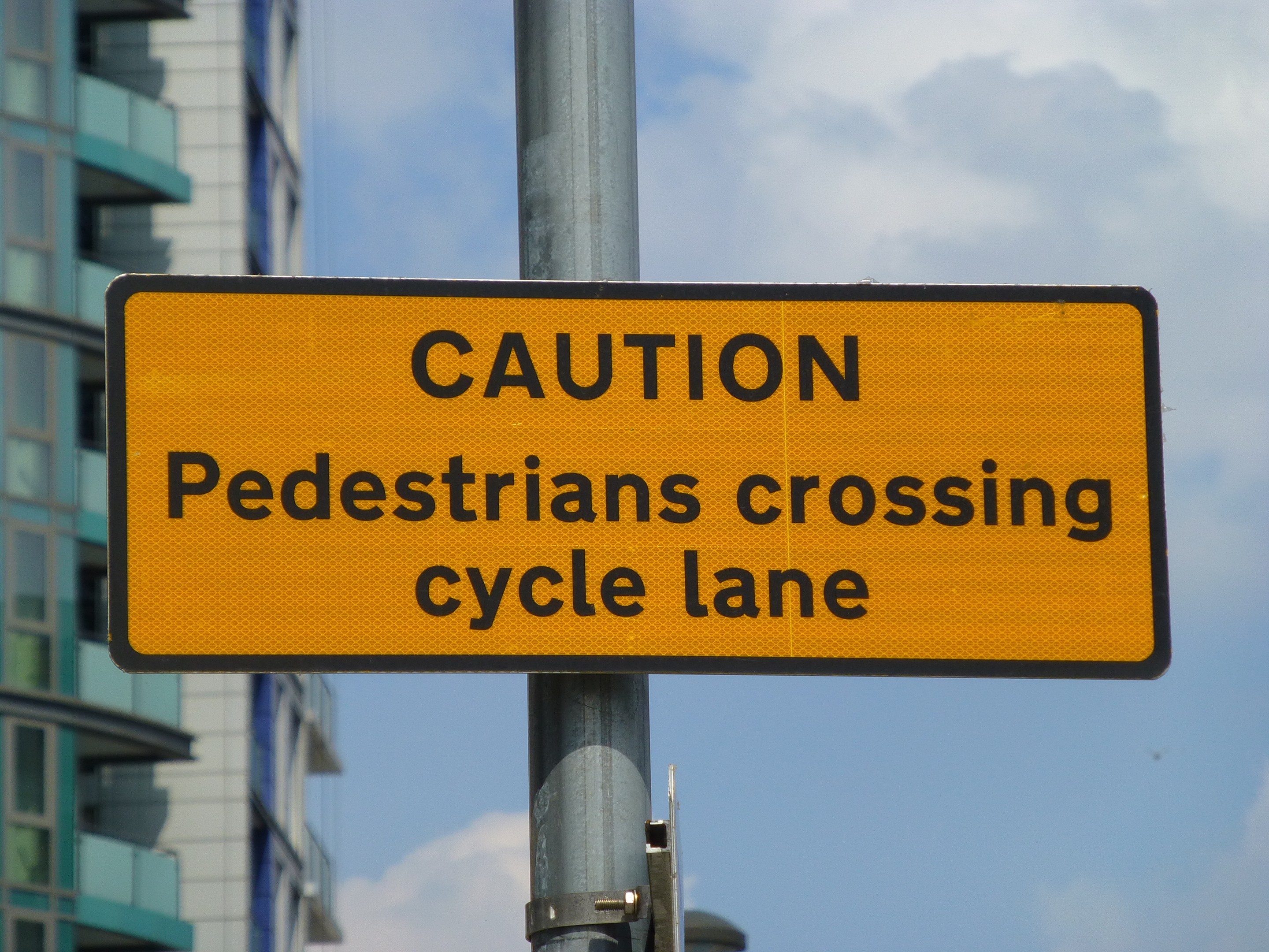 Gelbes Warnschild an einem Pfahl vor einem Gebäude, mit einem klaren blauen Himmel im Hintergrund, das 'Fußgänger überqueren Fahrradweg' anzeigt.