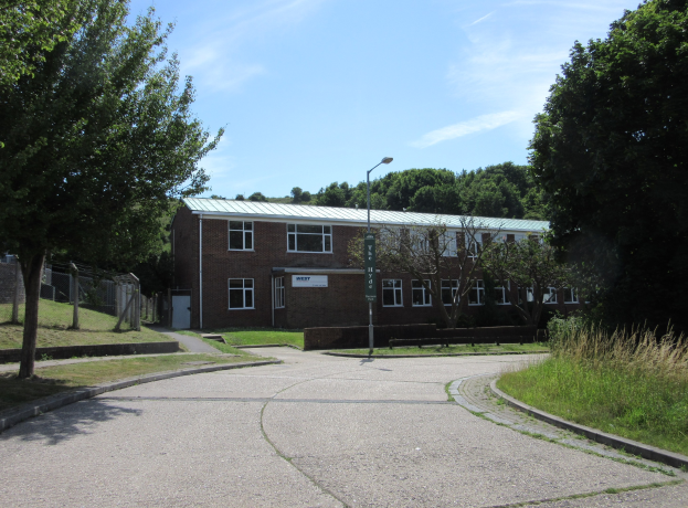 Ein Schulgebäude mit Fenstern, einem Straßenmast, einem Schild, einer Baumgruppe, Gras, einem Zaun und einem bewölkten Himmel, das sich in einem Park mit saftigem Gras befindet.