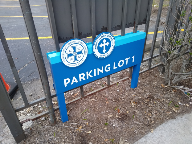 Ein blau-weißes Schild mit der Aufschrift 'Parkplatz 1' steht vor einem Metallzaun, im Hintergrund ist eine Straße zu sehen und rechts ein Baum.