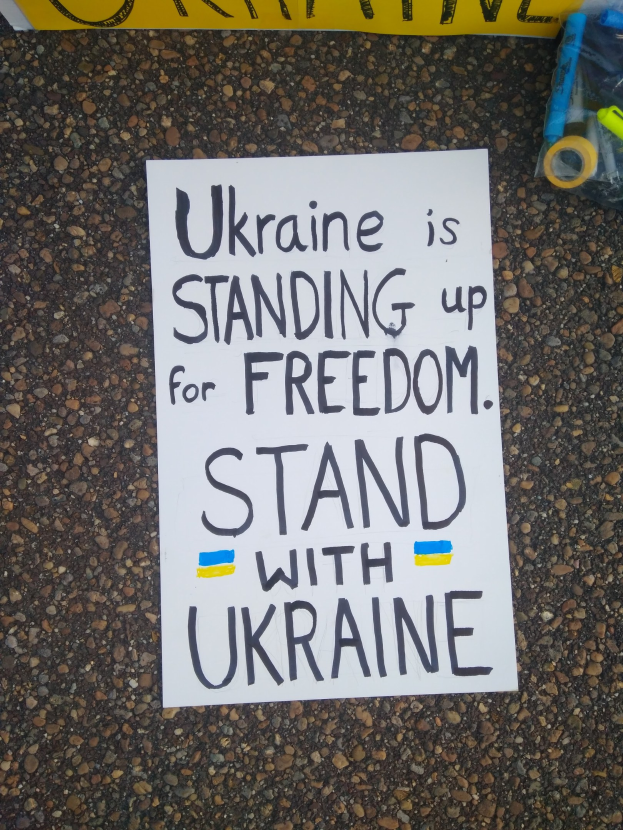 Ein weißes Blatt Papier mit dem Text "Ukraine steht für Freiheit auf, stellt Euch mit Ukraine" darauf, vor einem gelben Banner platziert, mit einer Plastikhülle mit verschiedenen Gegenständen rechts.