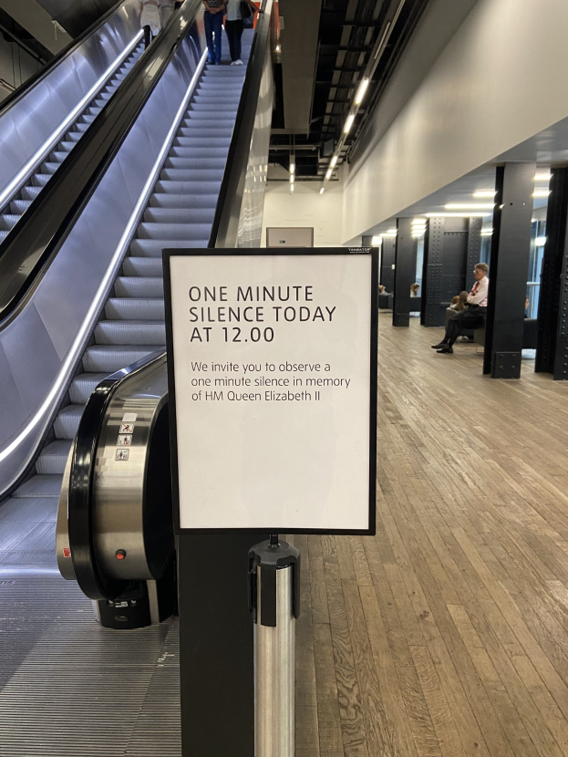 Eine Rolltreppe im Flughafen mit einem Schild, auf dem "Eine Minute Stille heute" steht, einige Menschen darauf und an der Decke angebrachte Lichter im Hintergrund.