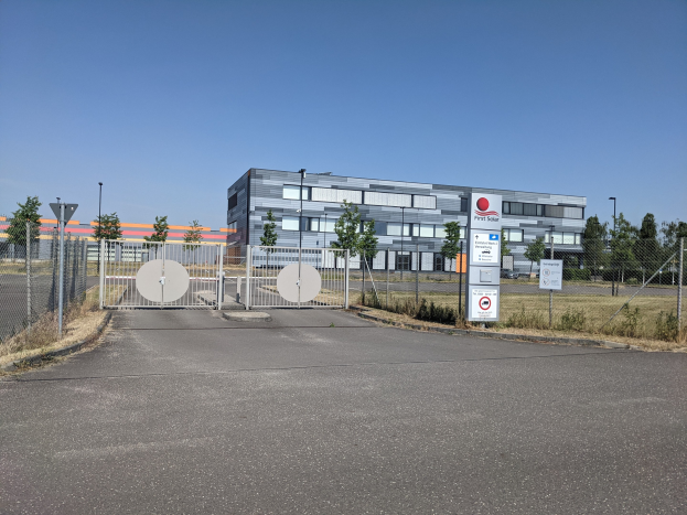 Großes industrielles Gebäude mit der Bezeichnung "First Solar" mit vielen Fenstern, umgeben von einem Metallzaun, umgeben von Bäumen, Pflanzen, Gras, Informationsschildern, Straßenmasten und einem Weg unter einem bewölkten Himmel.