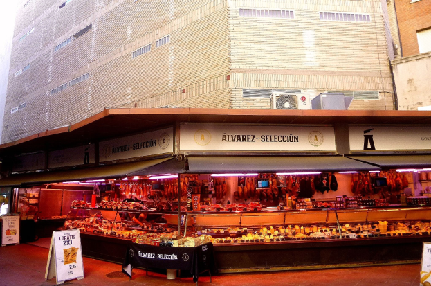 Ein belebter Markt mit verschiedenen Speisen, die unter Lichtern präsentiert werden, umgeben von Texttafeln und vor Buildings und einem klaren blauen Himmel.